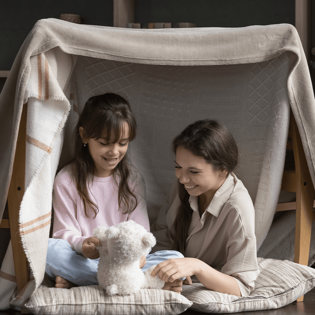 Mamma och dotter leker i en koja av filtar och stolar – roliga barnlekar inomhus.